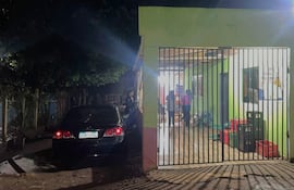 Entrada a un local con automóvil negro estacionado, dos mujeres conversan y un guardia observa desde el exterior.
