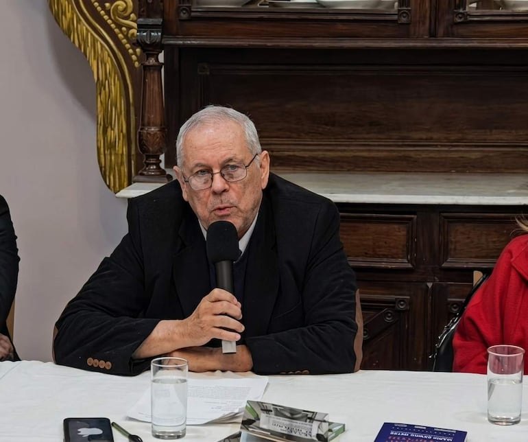 Mario Ramos Reyes, de edad avanzada, con abrigo oscuro y gafas, habla ante público mientras una mujer de abrigo rojo lo escucha.