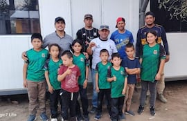 Grupo de diez personas, seis niños con camisetas verdes, hombres con camisetas de fútbol, sonrientes frente a estructura blanca.