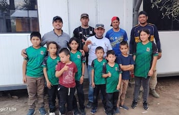 Grupo de diez personas, seis niños con camisetas verdes, hombres con camisetas de fútbol, sonrientes frente a estructura blanca.