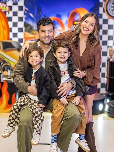 Hombre sostiene a niño mientras mujer sonriente acompaña. Fondo decorado con temática de carreras, ambiente colorido y festivo.
