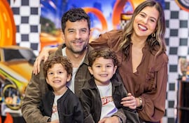 Hombre sostiene a niño mientras mujer sonriente acompaña. Fondo decorado con temática de carreras, ambiente colorido y festivo.