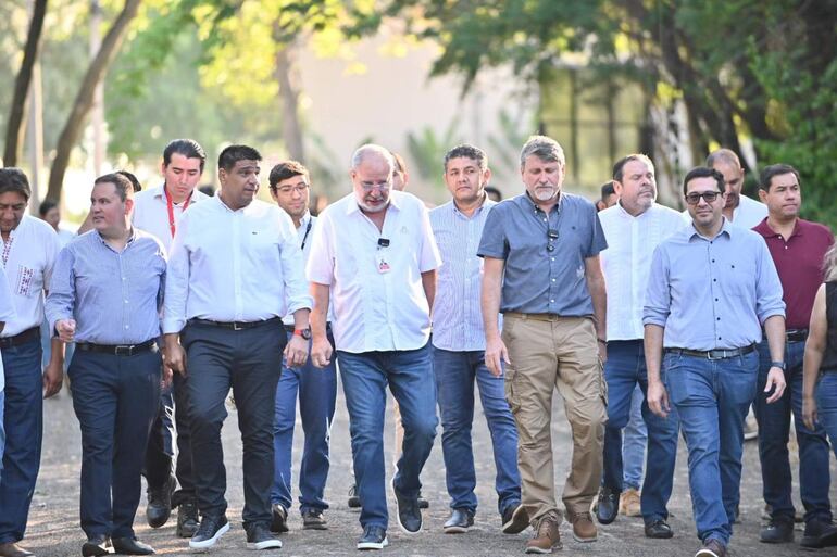 Denis Lichi en camiseta blanca rodeado de hombres con camisas y pantalones, caminando en un área verde con árboles.