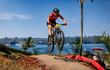 Ciclista masculino con camiseta roja y dorsal 87 salta sobre un obstáculo, rodeado de palmeras y paisaje acuático.