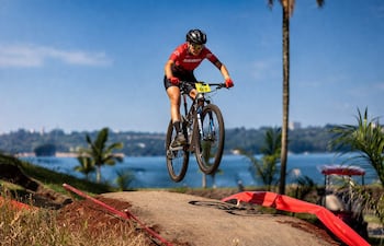 Ciclista masculino con camiseta roja y dorsal 87 salta sobre un obstáculo, rodeado de palmeras y paisaje acuático.