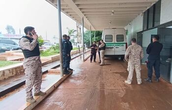 Hombre en uniforme militar hablando por teléfono junto a policías y personas en un entorno exterior húmedo conambulancia en el fondo.
