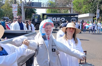 Hombres y mujeres con camisetas blancas y banderas participan en una manifestación, destacando un muñeco y una sombrero grande.