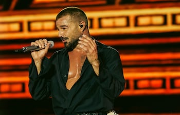 Ricky Martin canta en el escenario con micrófono, luciendo camisa negra y gestos apasionados, en un ambiente iluminado con tonos cálidos.