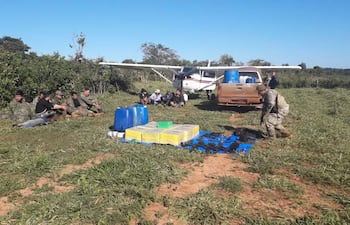 Grupo de policías con ropa de camuflaje y un hombre en uniforme militar junto a una avioneta y barriles en área verde.
