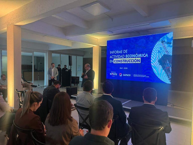 Orador en traje habla frente a público atento en una sala de conferencias moderna, con pantalla que muestra informe de construcción.