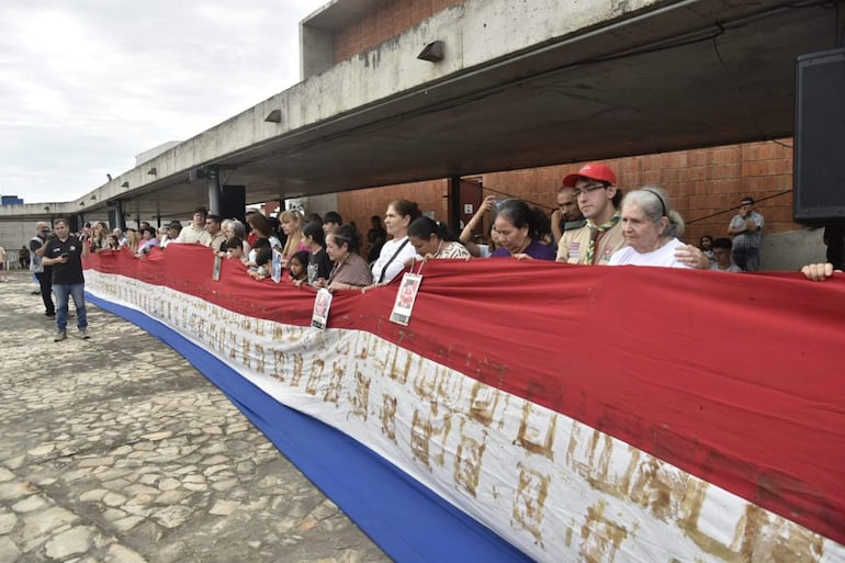 Aproximadamente 30 personas, vestidas con camisas neutras, sostienen un gran lienzo tricolor y fotos de seres queridos en un evento emotivo.