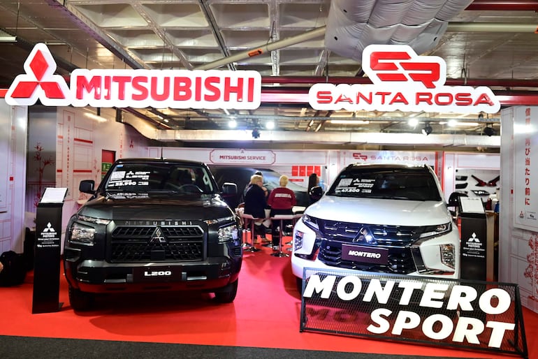 Stand con dos camionetas Mitsubishi L200 negra y Montero Sport blanca, rodeadas de personas conversando en un ambiente interior.