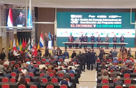 Panel formal con hombres y una mujer en trajes, prestando atención a orador en mesa. Público toma notas, banderas visibles.