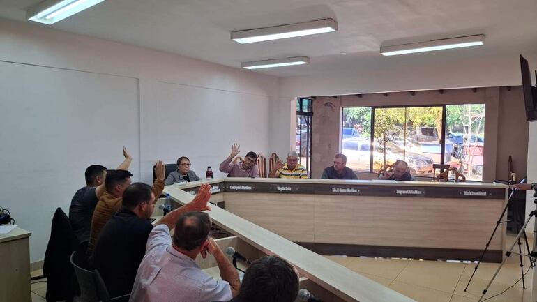 Reunión formal con siete participantes, dos hombres de camisa clara en el centro y cuatro levantan la mano en señal de voto.