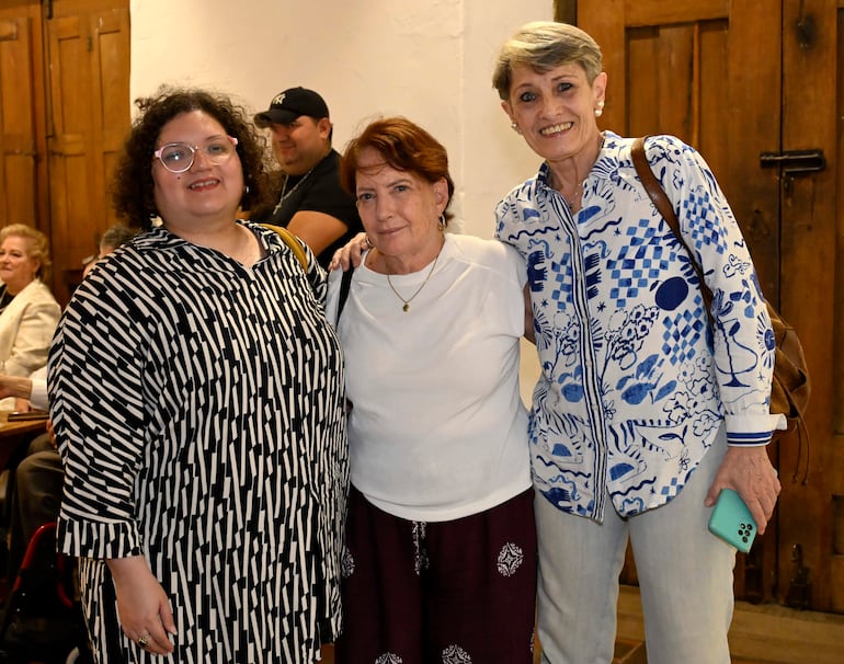 Tres mujeres sonrientes: una con camisa de rayas, otra con camiseta blanca y una más en blusa azul, en un ambiente cálido.