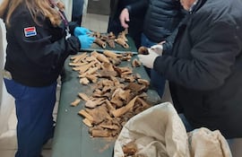 Mujer inspecciona piezas en mesa, usando guantes y mascarilla, mientras cuatro hombres discuten en un ambiente institucional.