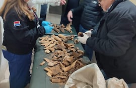 Mujer inspecciona piezas en mesa, usando guantes y mascarilla, mientras cuatro hombres discuten en un ambiente institucional.