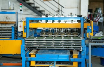 Interior de fábrica con máquina metálica azul y amarilla, sin personas presentes y piezas en proceso sobre la mesa.