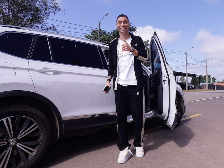 Miguel Almirón sonríe junto a su coche blanco, vistiendo un conjunto deportivo negro y blanco con el logo de Casablanca.