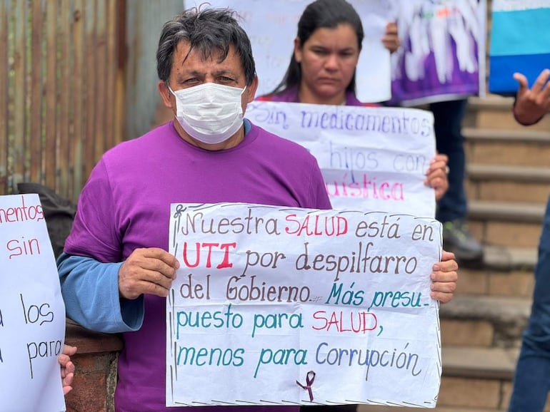 Hombre con camiseta morada y mascarilla sostiene cartel sobre salud pública mientras otros participantes observan detrás en una manifestación.