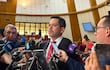 Raúl Latorre en saco negro y corbata roja, rodeado de periodistas, en un recinto legislativo con sillas rojas.