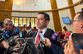 Raúl Latorre en saco negro y corbata roja, rodeado de periodistas, en un recinto legislativo con sillas rojas.