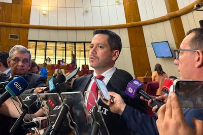 Raúl Latorre en saco negro y corbata roja, rodeado de periodistas, en un recinto legislativo con sillas rojas.