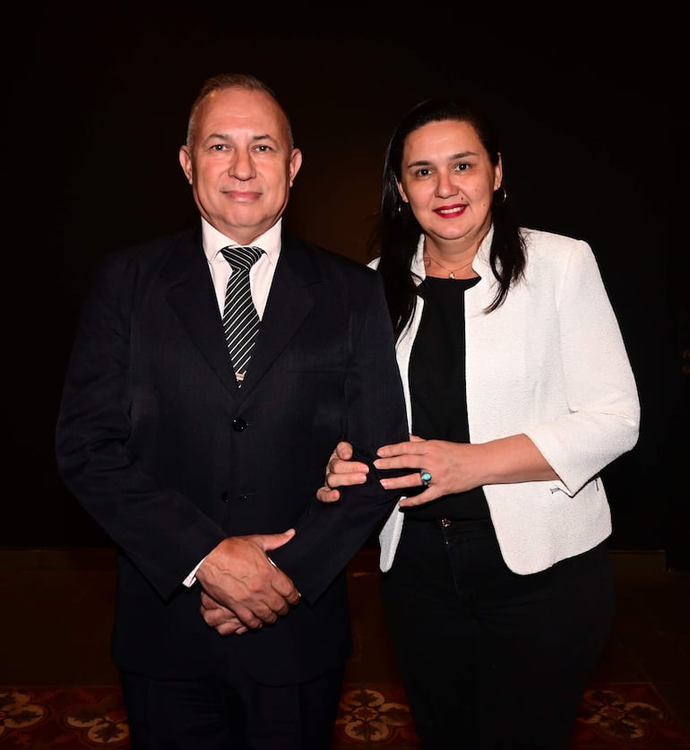Embajador de Francia en traje oscuro y mujer en blusa negra y abrigo blanco, posando juntos en un ambiente formal con fondo oscuro.