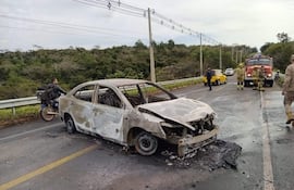 Hombres en trajes de bomberos evalúan automóvil quemado en la carretera, con vegetación densa al fondo y tráfico parcial.