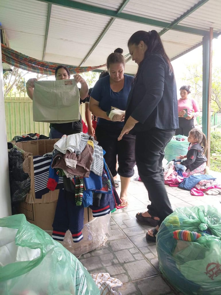 Cinco mujeres en ambiente iluminado clasifican ropa donada, mientras una niña juega a su lado.