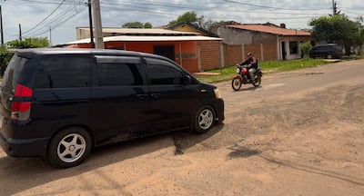 Un coche oscuro estacionado, con una motocicleta roja avanzando y casas de ladrillo en un día nublado.