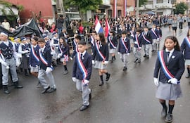 Niños marchan en uniforme oscuro con cintas en blanco, azul y rojo, sosteniendo banderas, en un ambiente festivo.