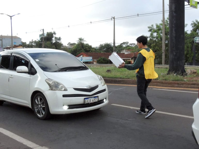 Joven con chaleco amarillo sostiene una caja, caminando hacia un automóvil blanco en un entorno urbano parcialmente nublado.
