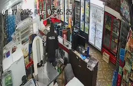 Agente de policía con chaqueta que dice 'POLICÍA' revisa detrás del mostrador en un comercio con estanterías llenas de productos.