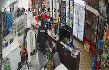 Agente de policía con chaqueta que dice 'POLICÍA' revisa detrás del mostrador en un comercio con estanterías llenas de productos.
