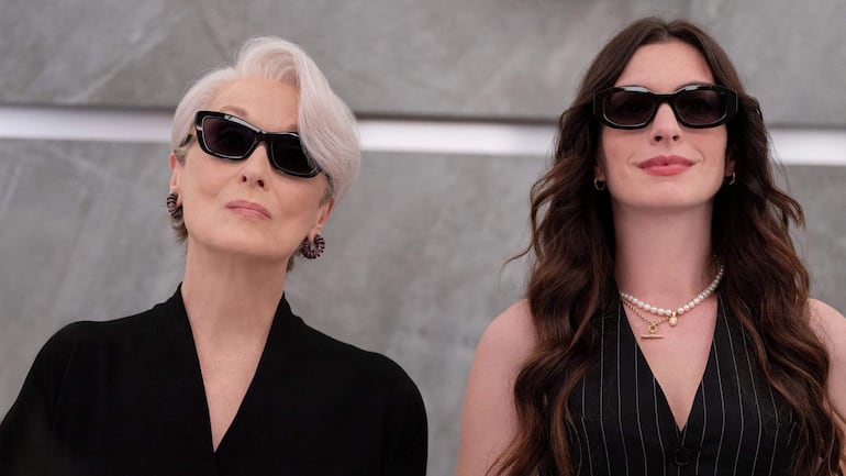 Anne Hathaway con cabello corto y prenda negra, y Meryl Streep con blusa a rayas y collar de perlas, ambas con gafas oscuras.