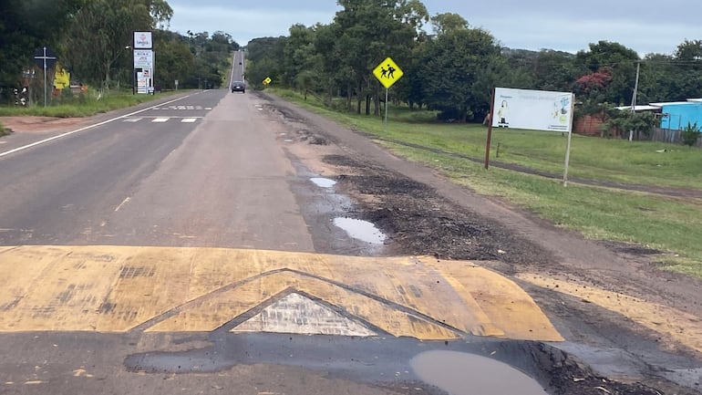 Un vehículo se aproxima por la ruta deteriorada, con señales de advertencia y un reductor de velocidad visible en un día nublado.