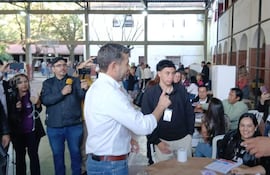 Pedro Alliana en camisa blanca conversando con un joven en un recinto de votación, con varios periodistas alrededor.