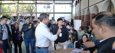Pedro Alliana en camisa blanca conversando con un joven en un recinto de votación, con varios periodistas alrededor.