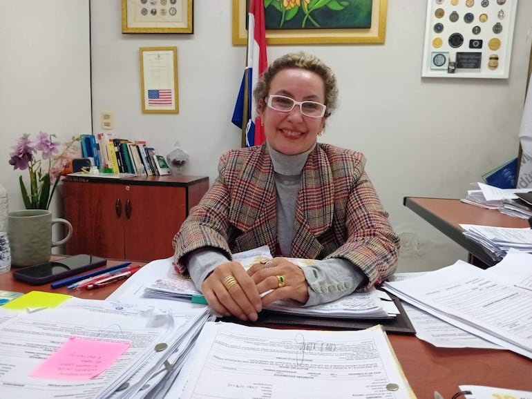 Irma Llano, con cabello corto y gafas, revisa papeles en su oficina rodeada de libros y decoraciones mientras sonríe.