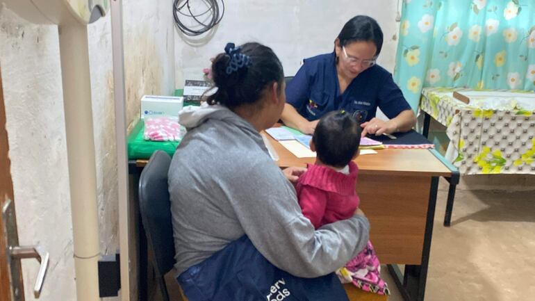 Mujer con niño en brazos y profesional de salud en escritorio, rodeados de documentos y un ambiente interior claro.