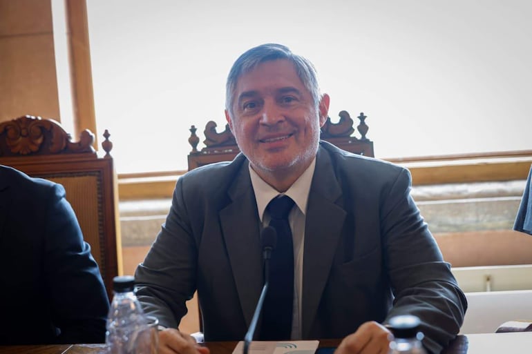 Santiago Peña sonríe en traje gris con corbata negra, sentado en mesa durante reunión, mientras otro hombre parcialmente visible lo acompaña.