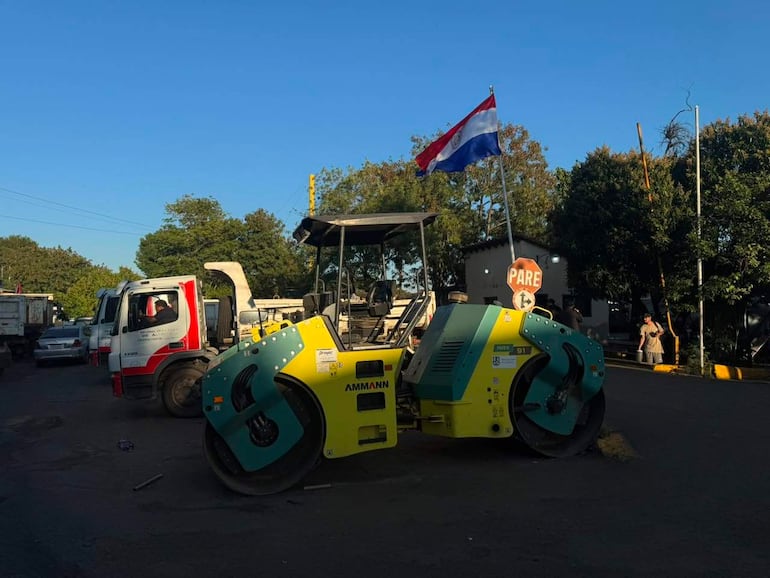 Máquina rodillo amarilla y verde en obra de construcción, con bandera paraguaya, y cartel de stop visible en el entorno.