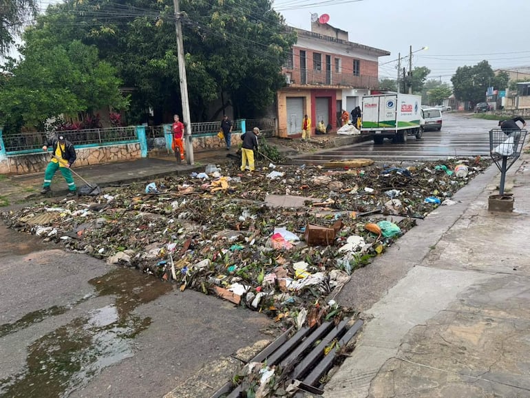 Seis operarios con chalecos amarillos y verdes recogen basura en un suelo mojado, junto a un camión de recolección en un ambiente descuidado.