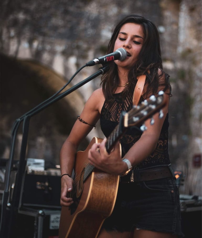 Kelly Ou Moi, de cabello largo y blusa de encaje negro, canta y toca la guitarra con mirada concentrada en un ambiente desenfocado.