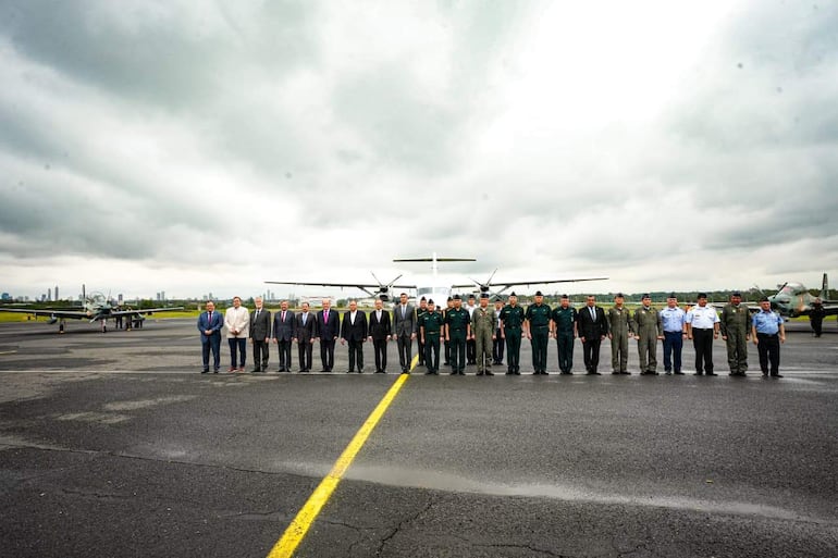Un grupo de 20 personas en trajes formales y uniformes militares alineados en el aeropuerto, mostrando expresiones felices.