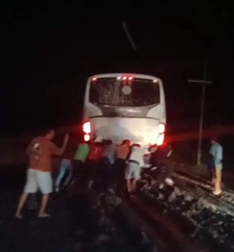Diez personas empujan un autobús blanco atascado en un camino de tierra, iluminadas por las luces traseras del vehículo.