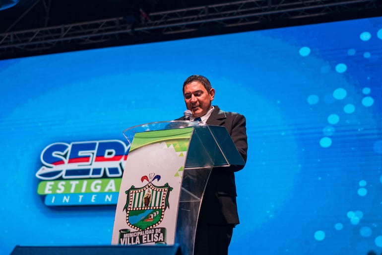 Hombre en traje oscuro se inclina hacia un micrófono mientras sostiene un documento en un evento con fondo decorativo.