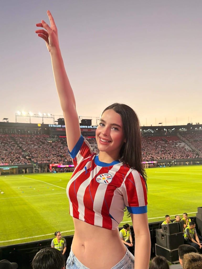 Mujer joven con camiseta rayada roja y blanca sonríe en un estadio lleno, levantando un brazo con entusiasmo.