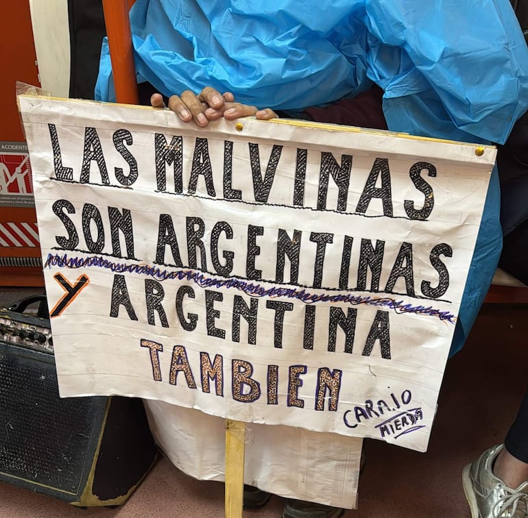 Protestante con poncho azul sostiene un cartel que dice 'LAS MALVINAS SON ARGENTINAS', semblante decidido en un espacio público.